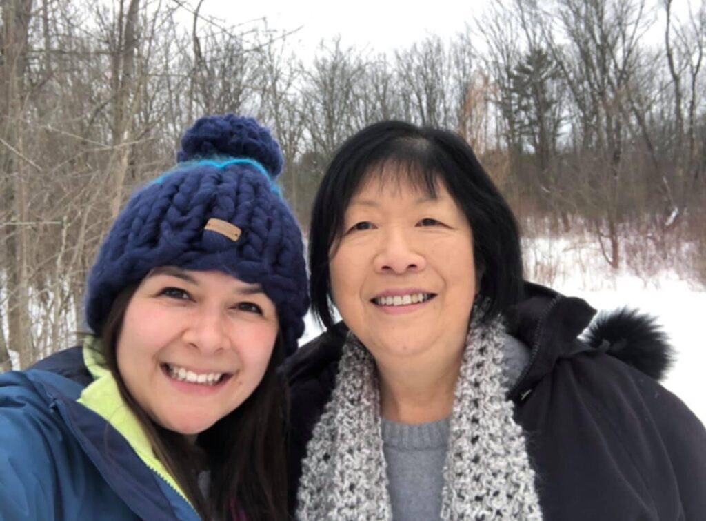 A selfie of two smiling women in winter attire against a snowy landscape. The younger woman on the left wears a knitted hat and the older woman on the right, her mother, wears a grey knit scarf and black winter jacket. Trees without leaves and covered in snow form the backgound. 