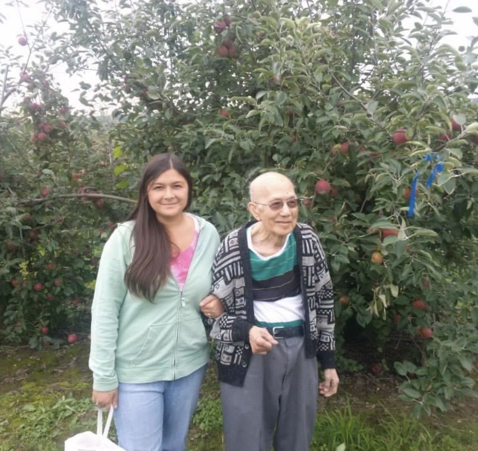 A young woman with long dark hair wearing a light green hoodie, stands next to her grandfather and holds his arm in an apple orchard. The elderly man has glasses and is dressed in a patterned shirt and sweater. Both are smiling and the background is filled with apple trees with many red apples.