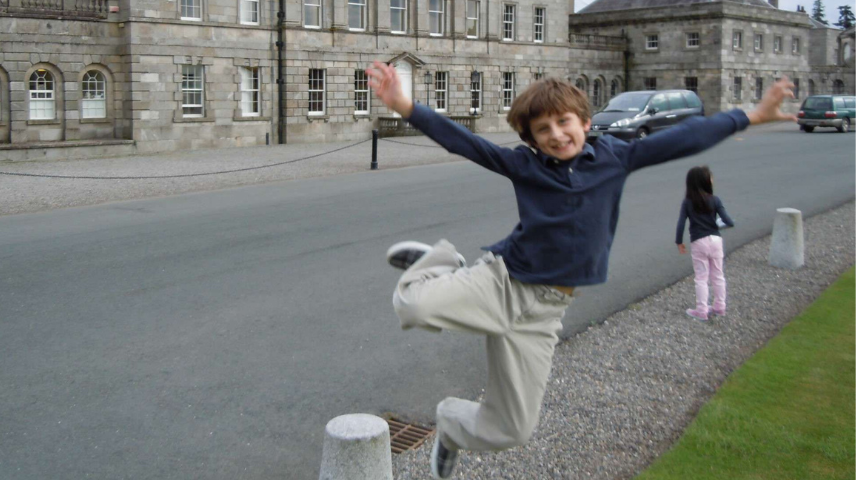 A joyful child with brown hair and blue jacket jumps mid-air with his arms spread wide, smiling broadly, in front of a large stone building.