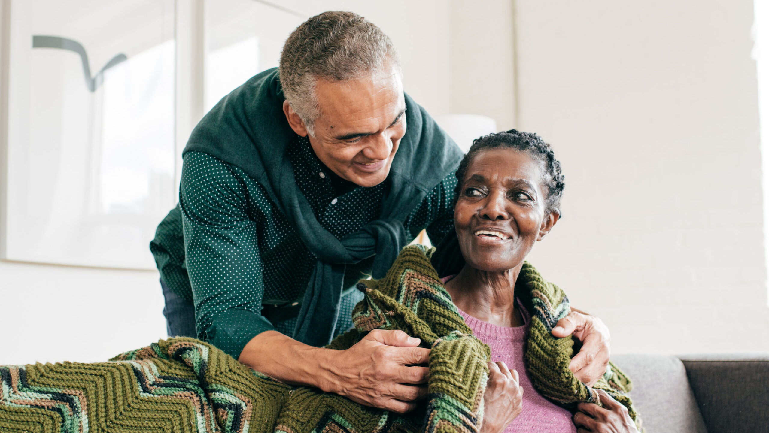 man standing behind elderly woman hugging her with a blanket