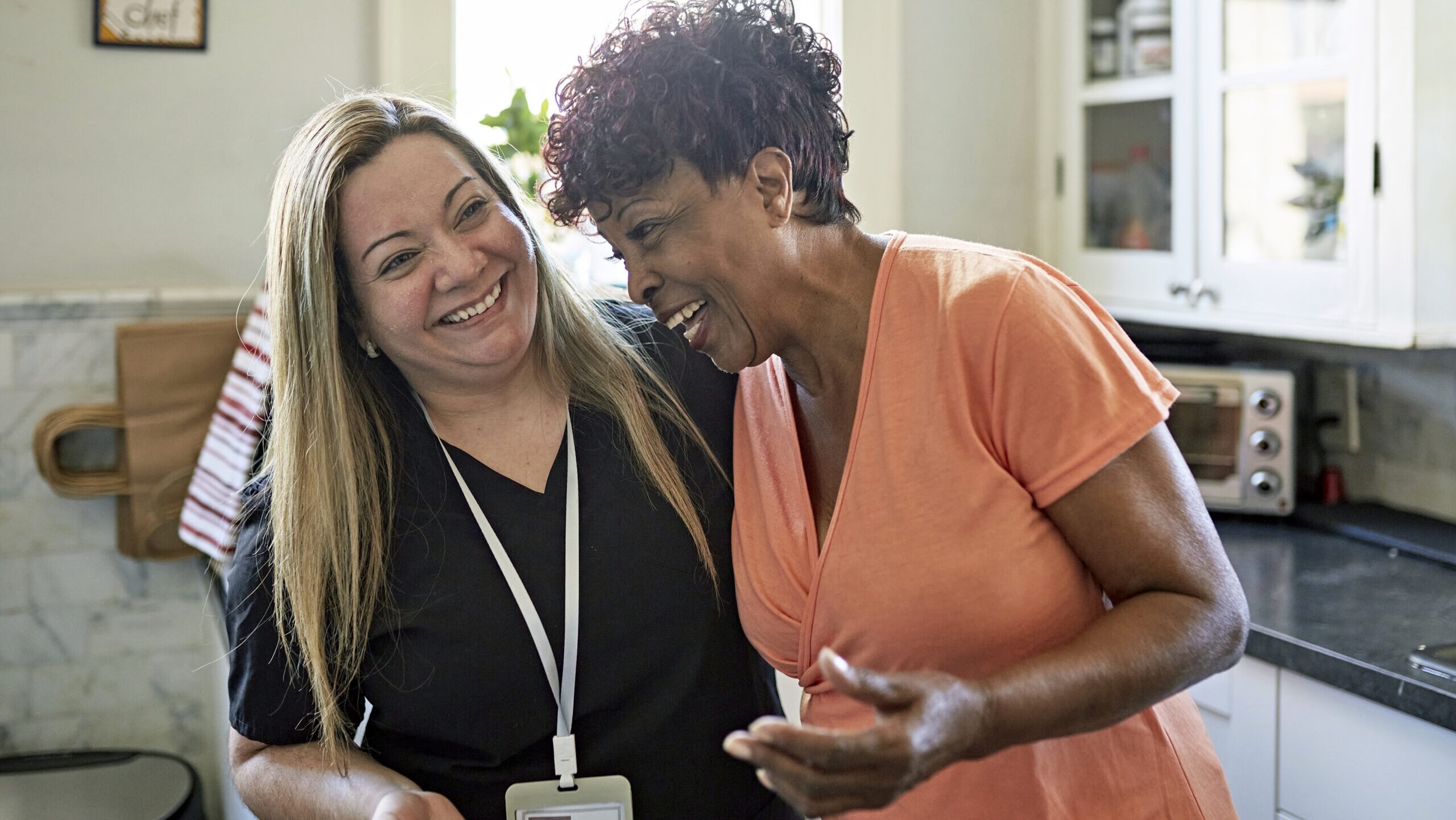 Woman and healthcare worker standing with arms around each other and laughing during routine house call.
