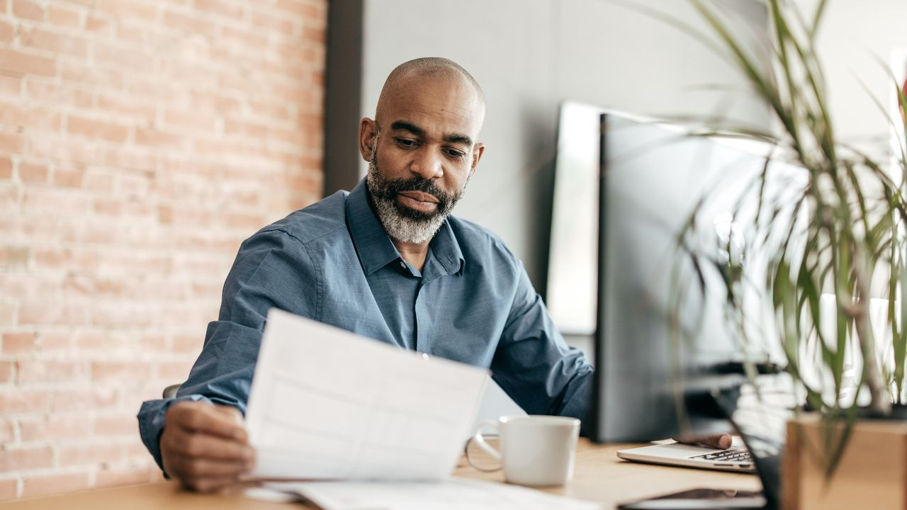 man in a blue denim shirt looks at a piece of paper while he sits in front of a computer
