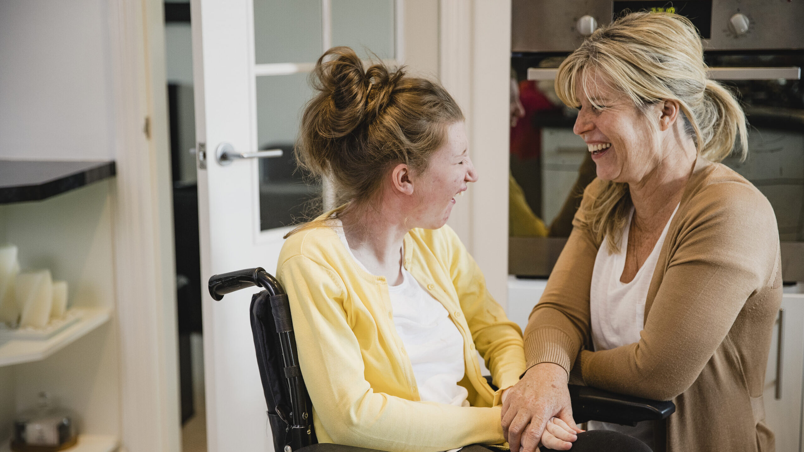 female participant in a wheelchair, wearing a yellow sweater, laughing with female caretaker who is kneeling