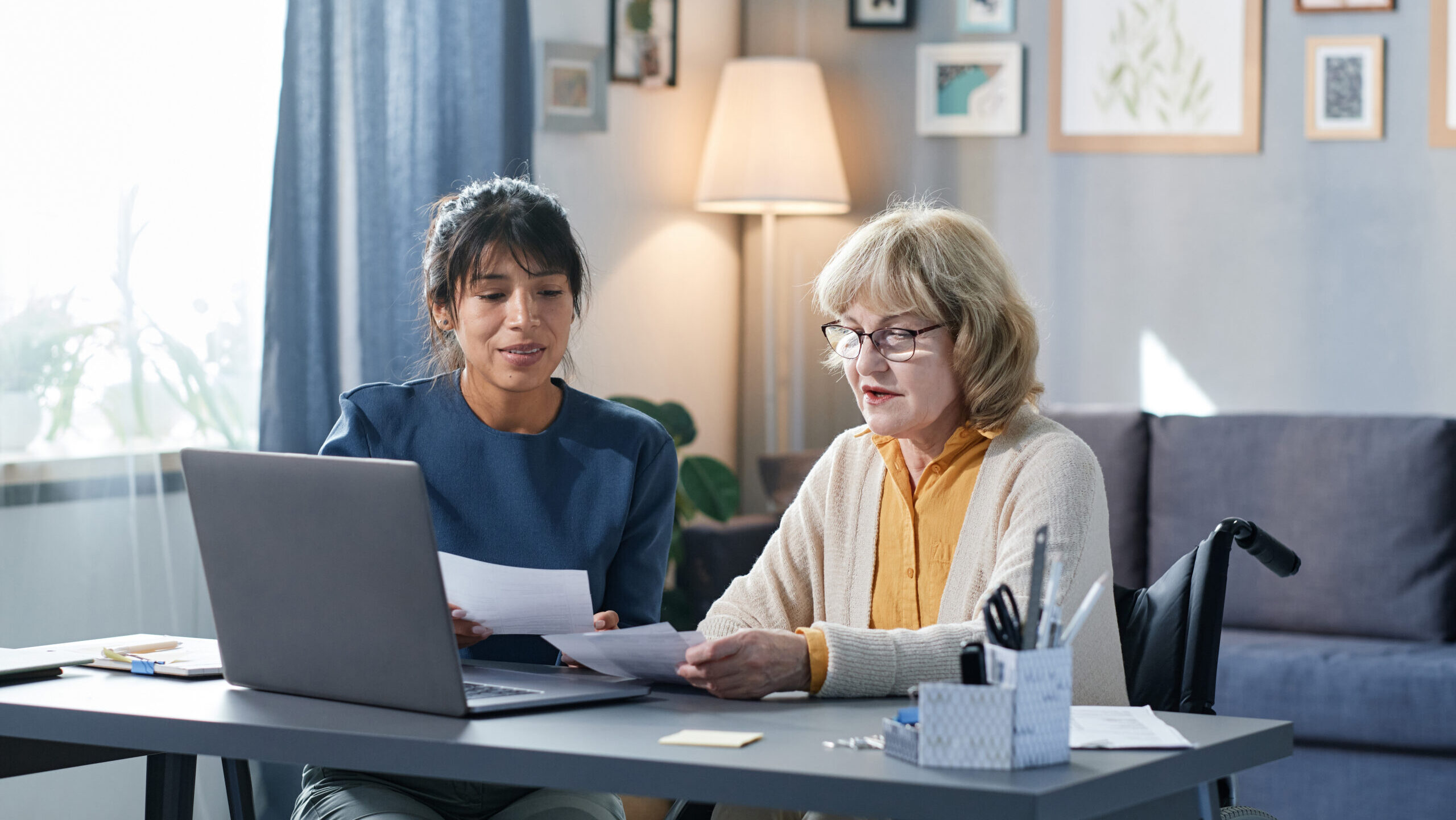 senior woman sitting in a wheel chair at a desk, being helped by a woman with dark brown hair. they are looking at papers together