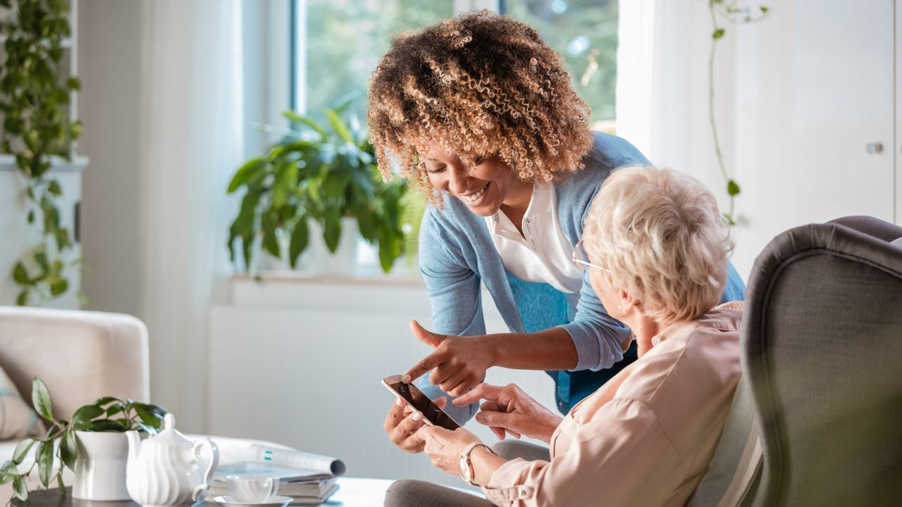 female caregiver assisting female, elderly participant who is seated. they are looking at a phone