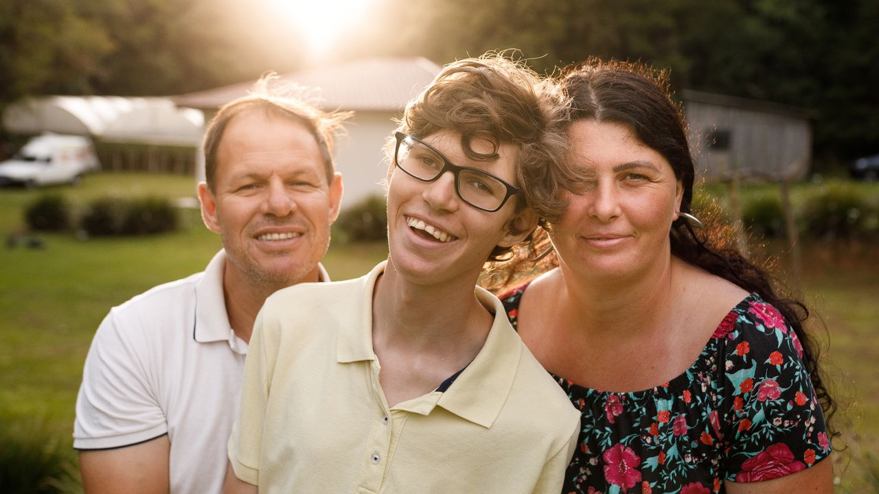 A smiling teenage boy stands between his two parents outdoors at sunset, representing a family supported through Pennsylvania's Participant-Directed Services.