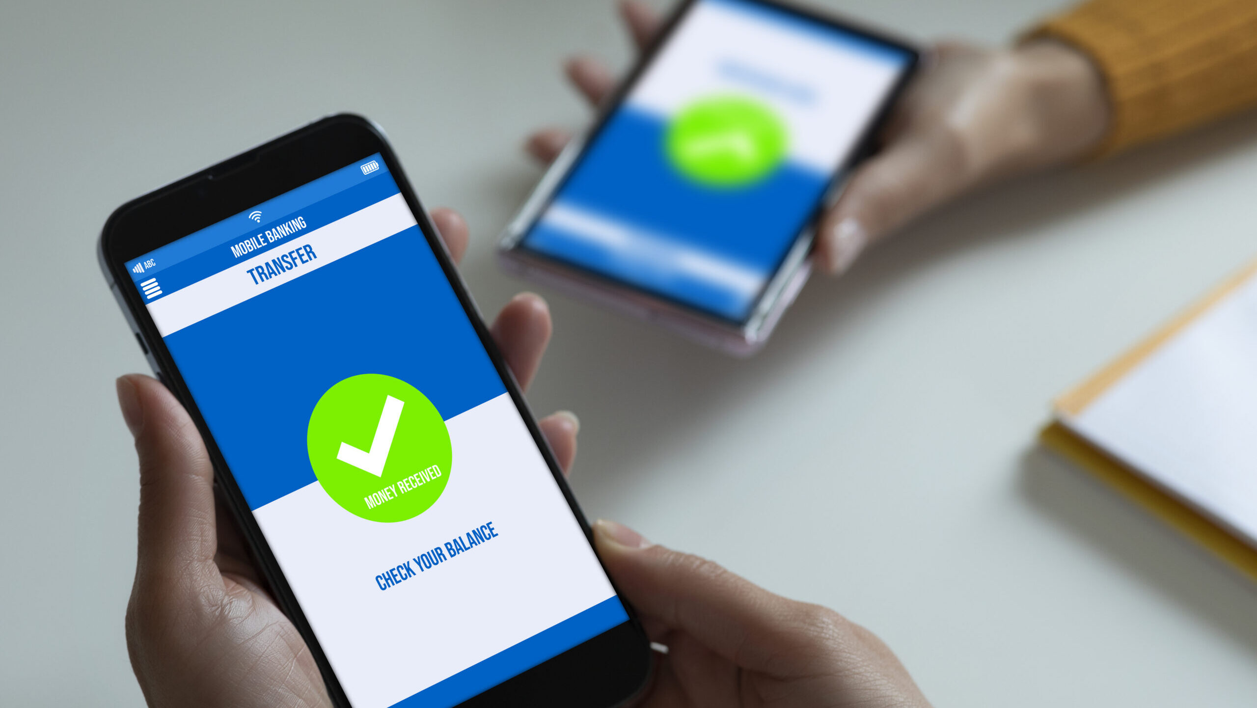 Close-up of a hand holding a smartphone showing a mobile banking transfer confirmation with a green checkmark and "Money Received," with a second phone displaying a similar screen blurred in the background.