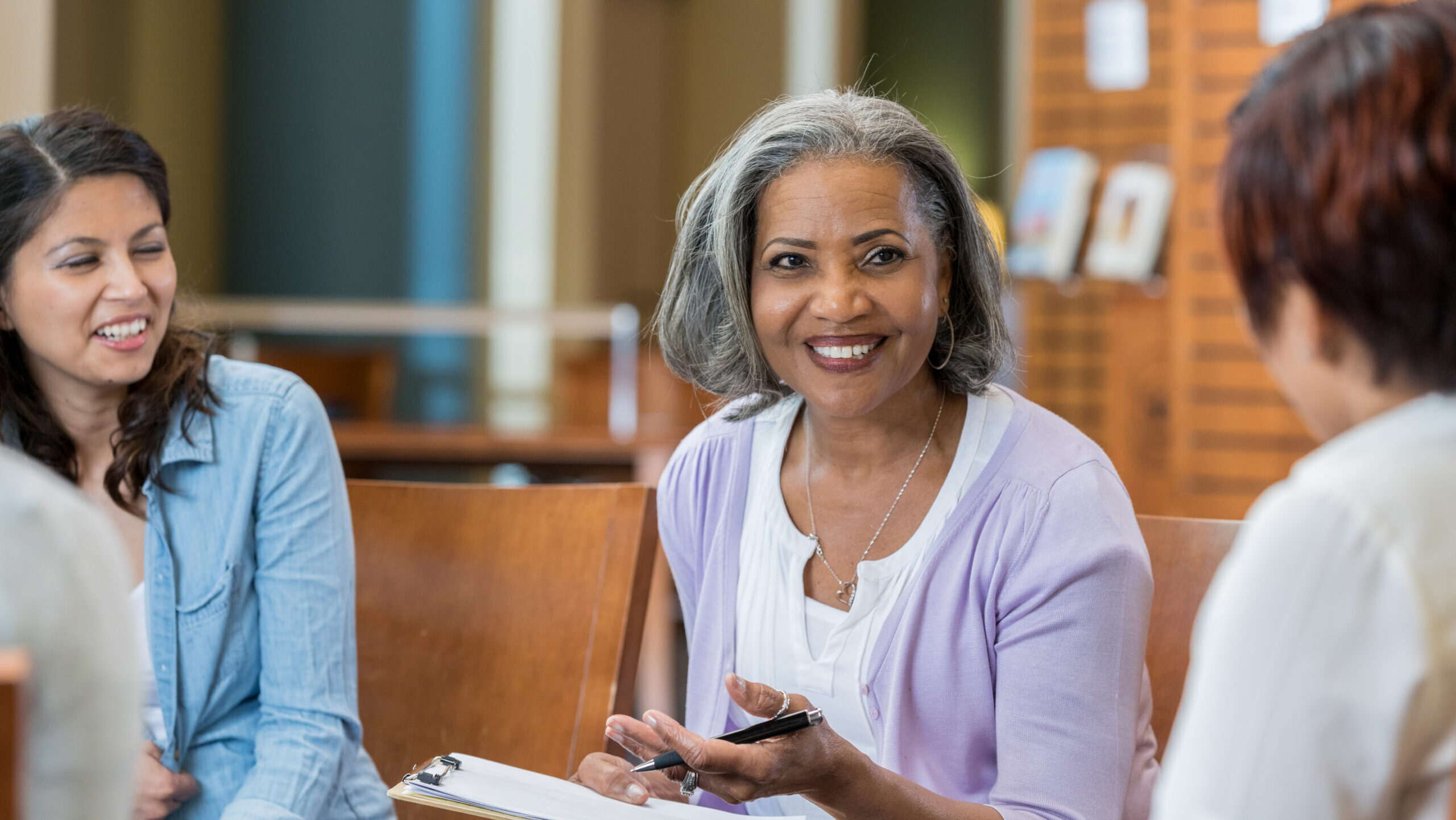A woman with gray hair holding a clipboard smiles while talking with two other women seated in a circle, appearing to lead a group discussion.