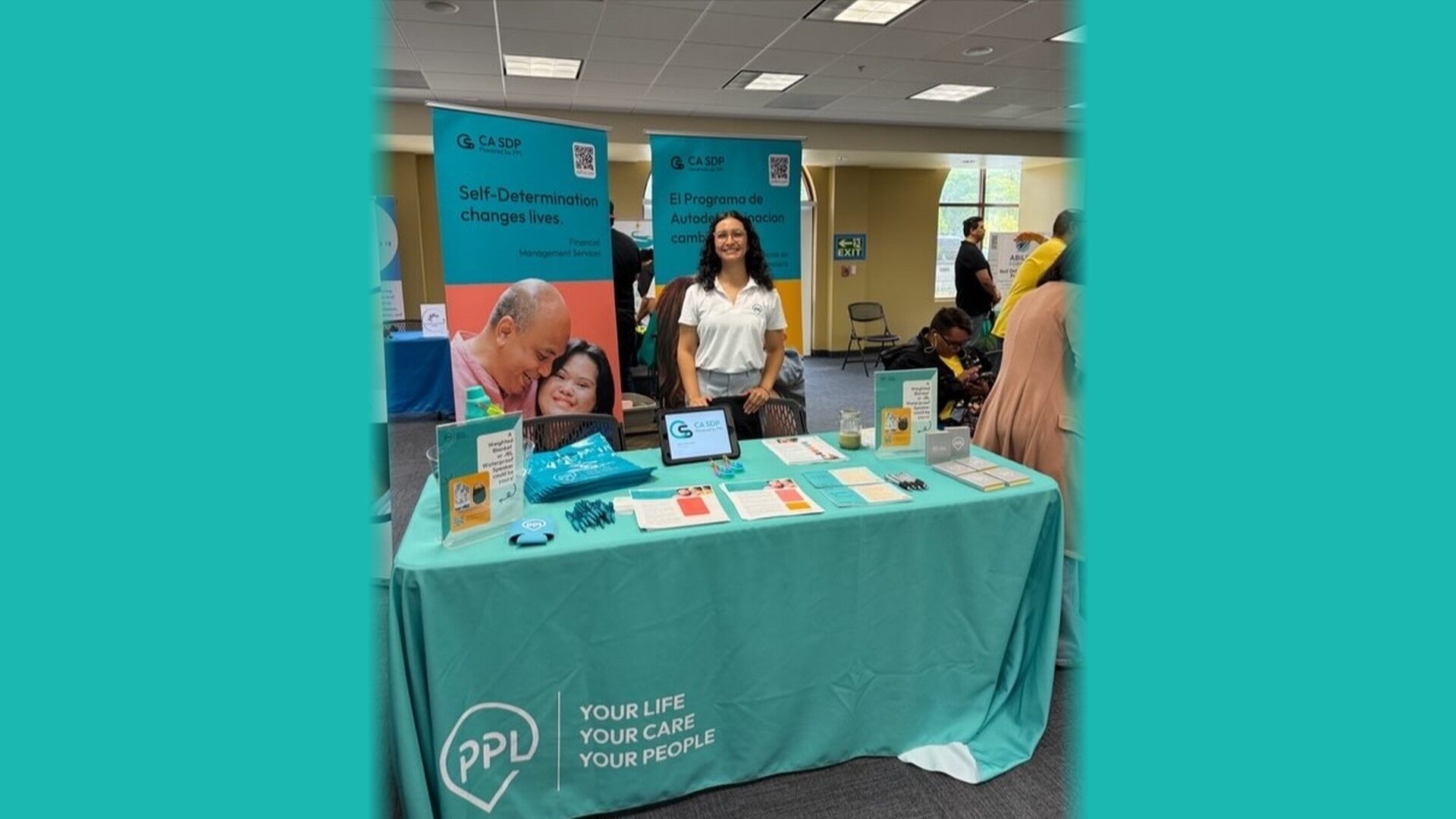 Tanya, with long, dark curly hair and glasses, smiles in a white polo behind our teal CA SDP table at a community event.