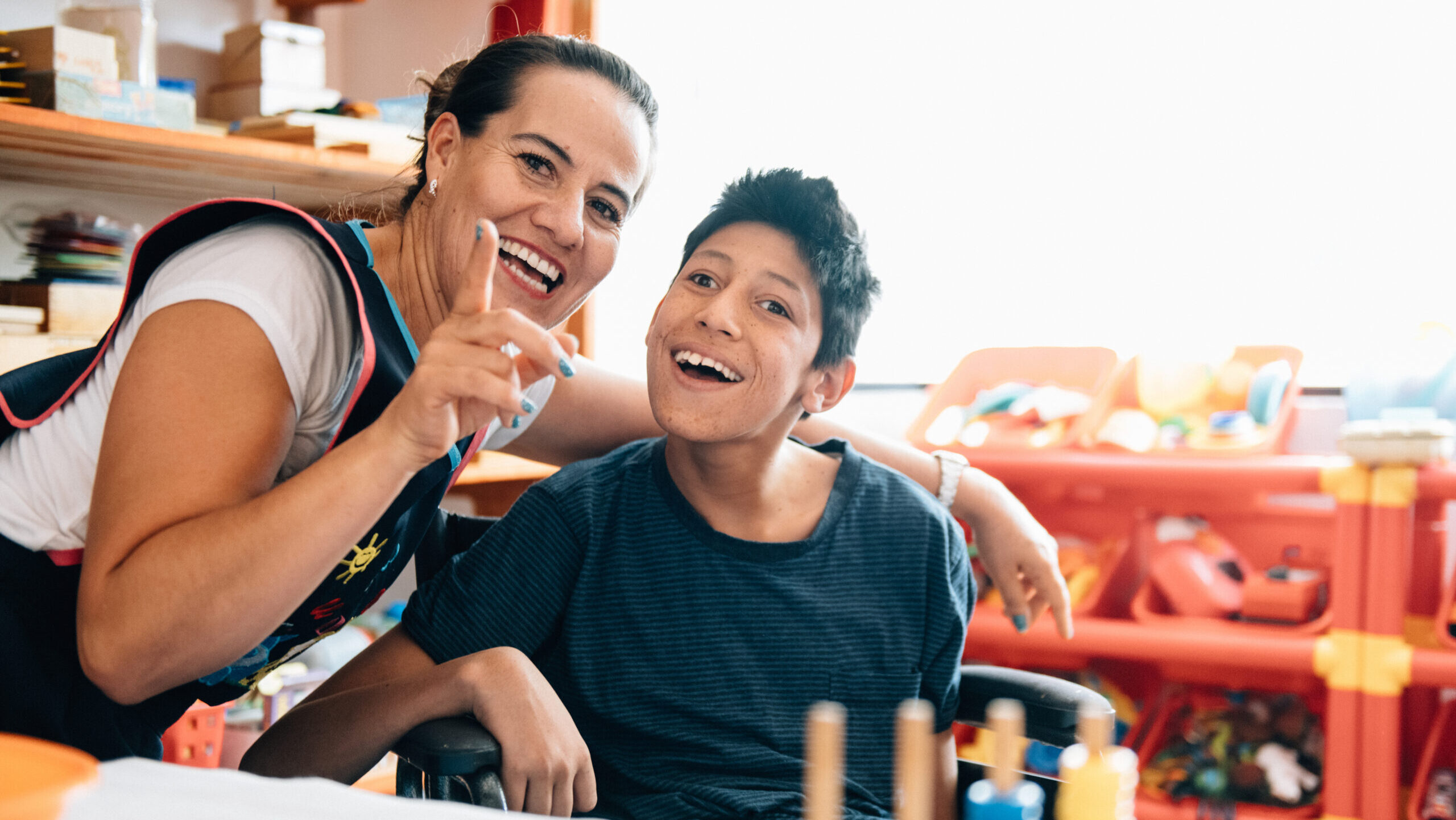 A smiling woman leans in beside a smiling teenager who uses a wheelchair, in a bright room filled with colorful toys and shelves.
