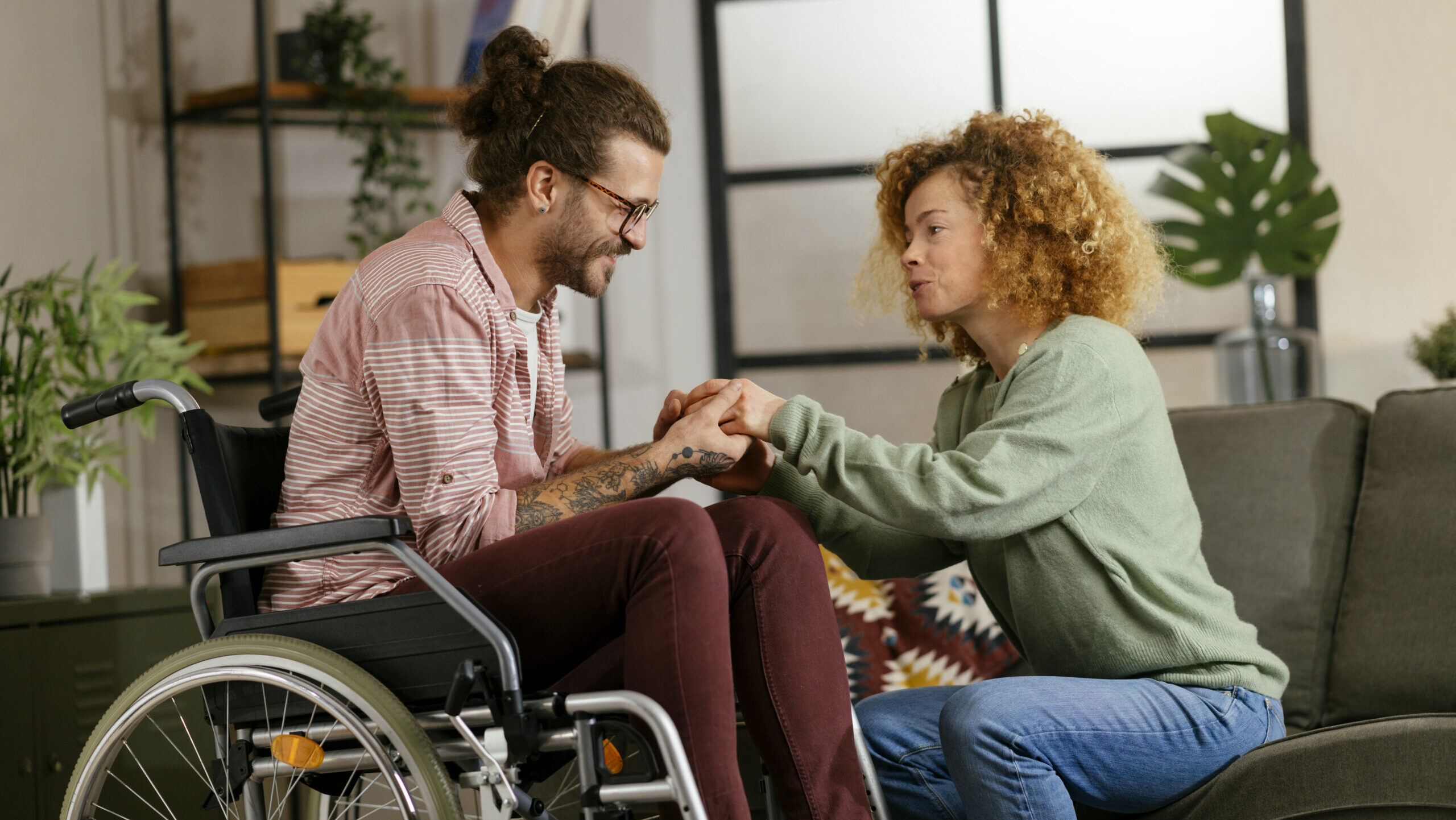 A man in a wheelchair and a woman sitting on a couch hold hands and talk in a living room.