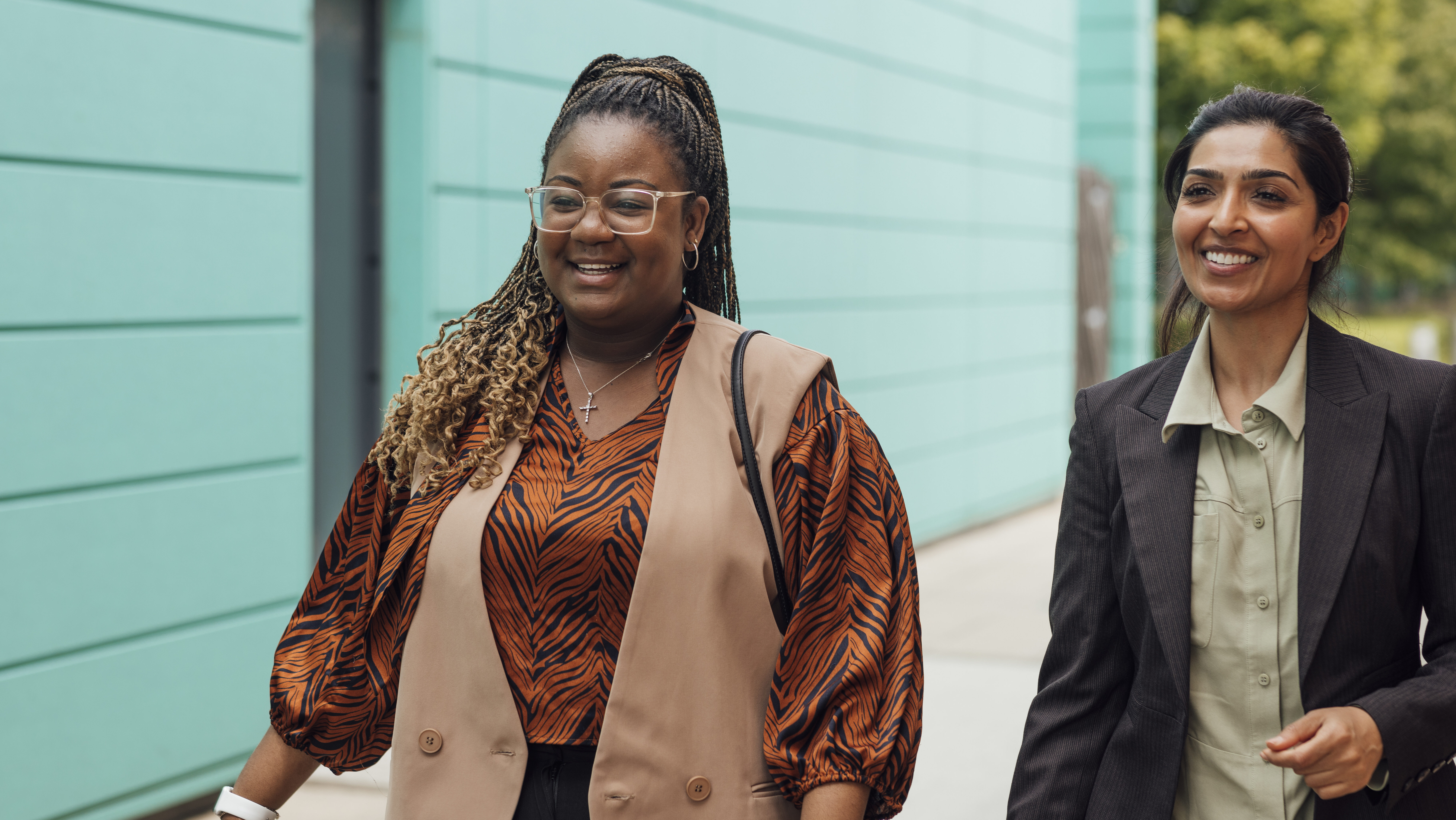Two smiling professional women walking together outside along a teal building.