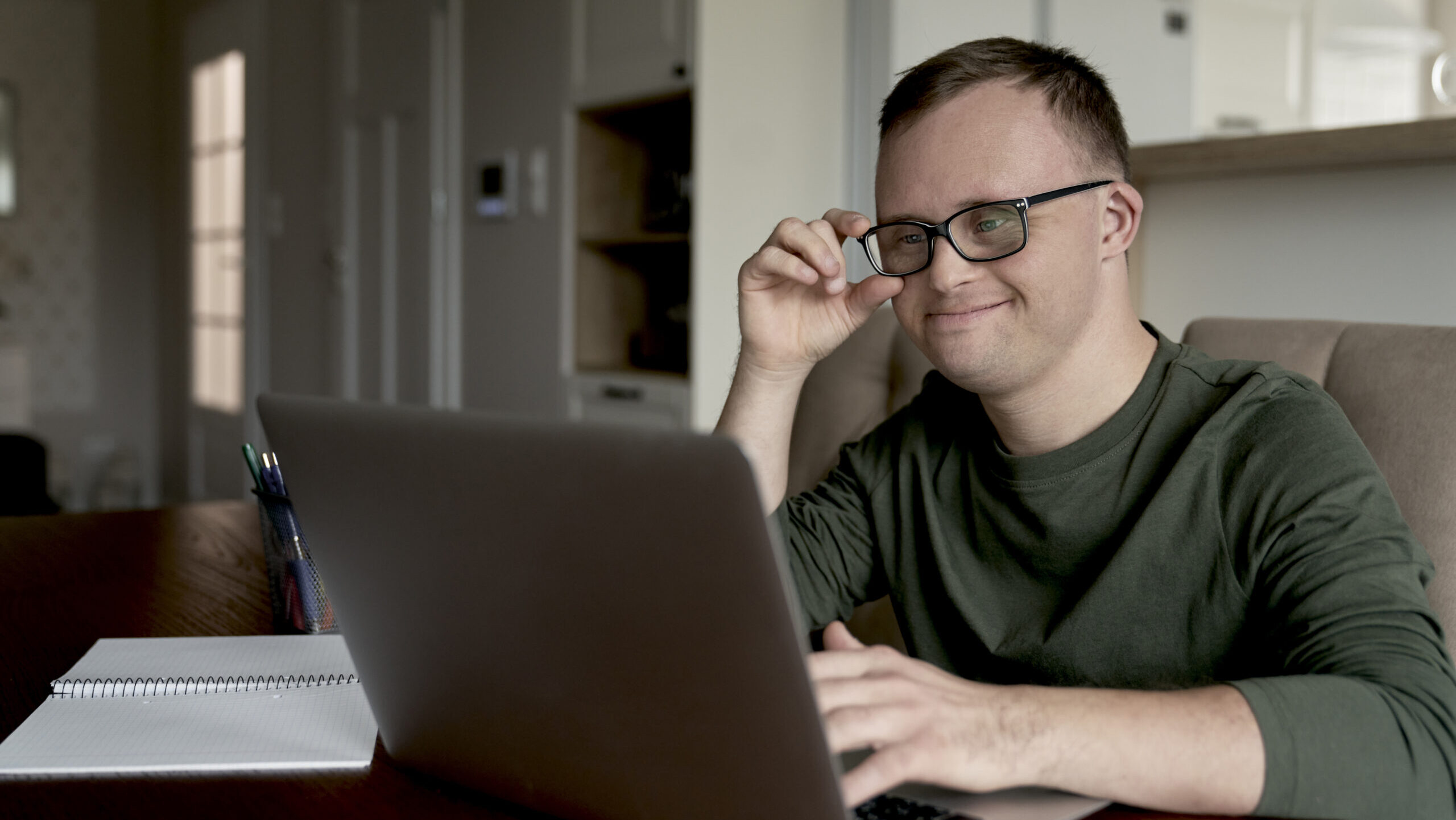 Man with Down syndrome wearing glasses and a green shirt, smiling while typing on a laptop at a kitchen table