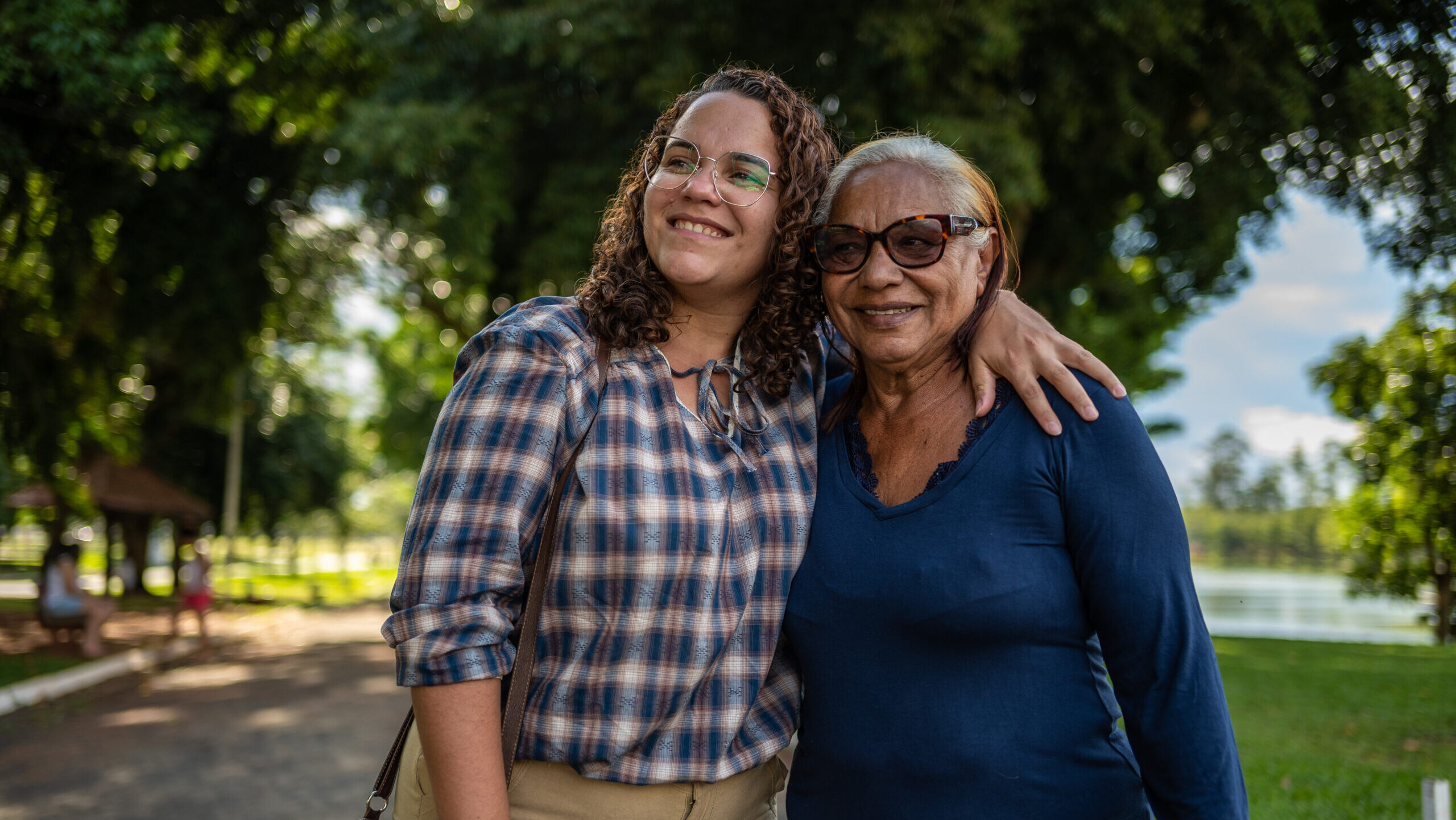 Two women smiling outdoors in a park, with the younger woman’s arm around the older woman’s shoulders and trees and a lake behind them.